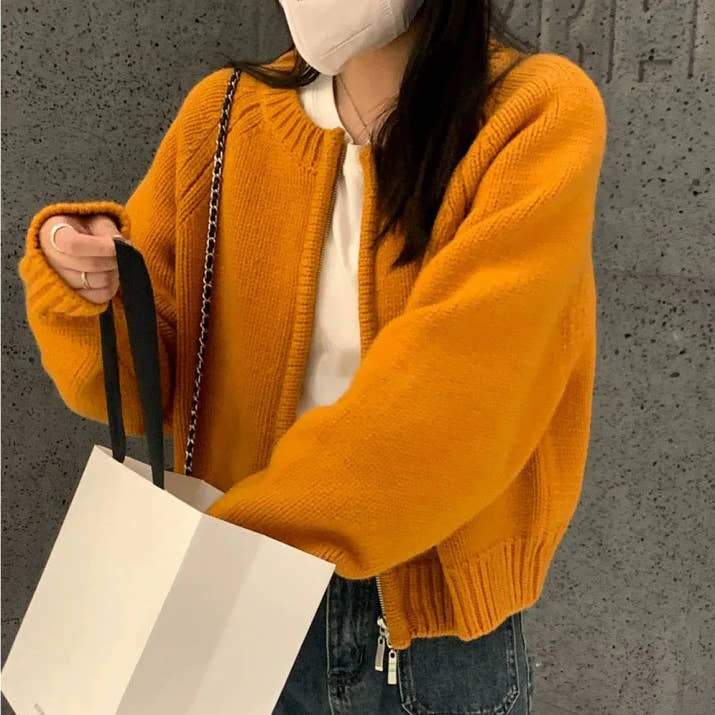 Person wearing a mustard yellow sweater holding a white bag against a gray wall.