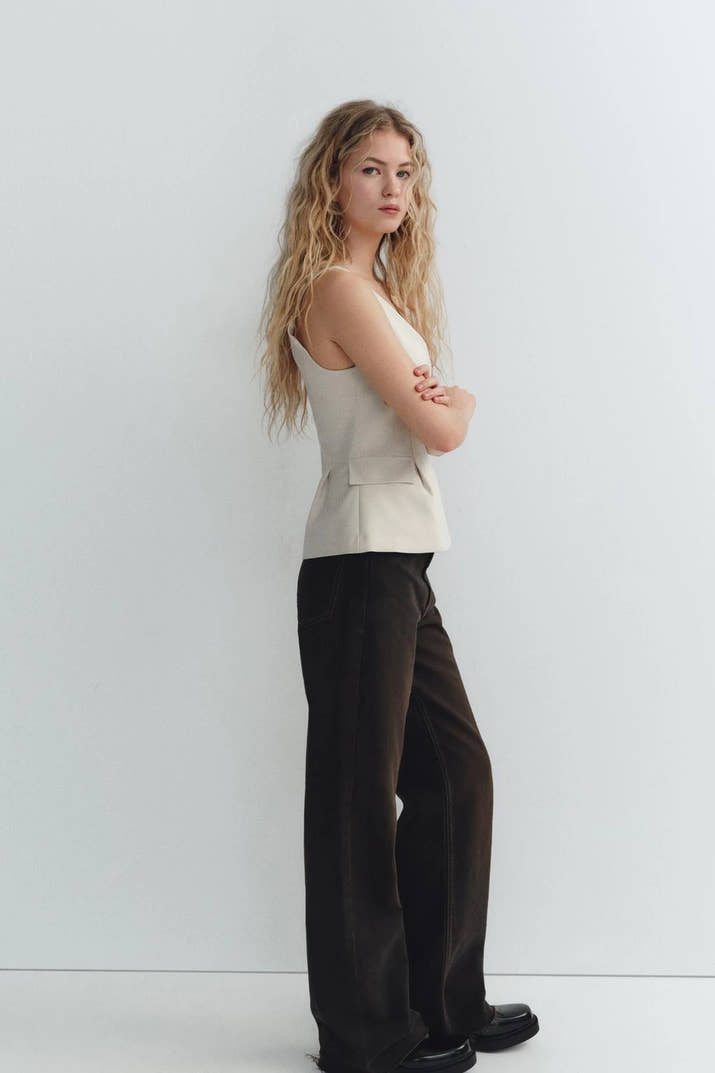 Woman wearing a beige sleeveless V-neck crop top with ruffled detail and zipper, styled casually in a café setting.