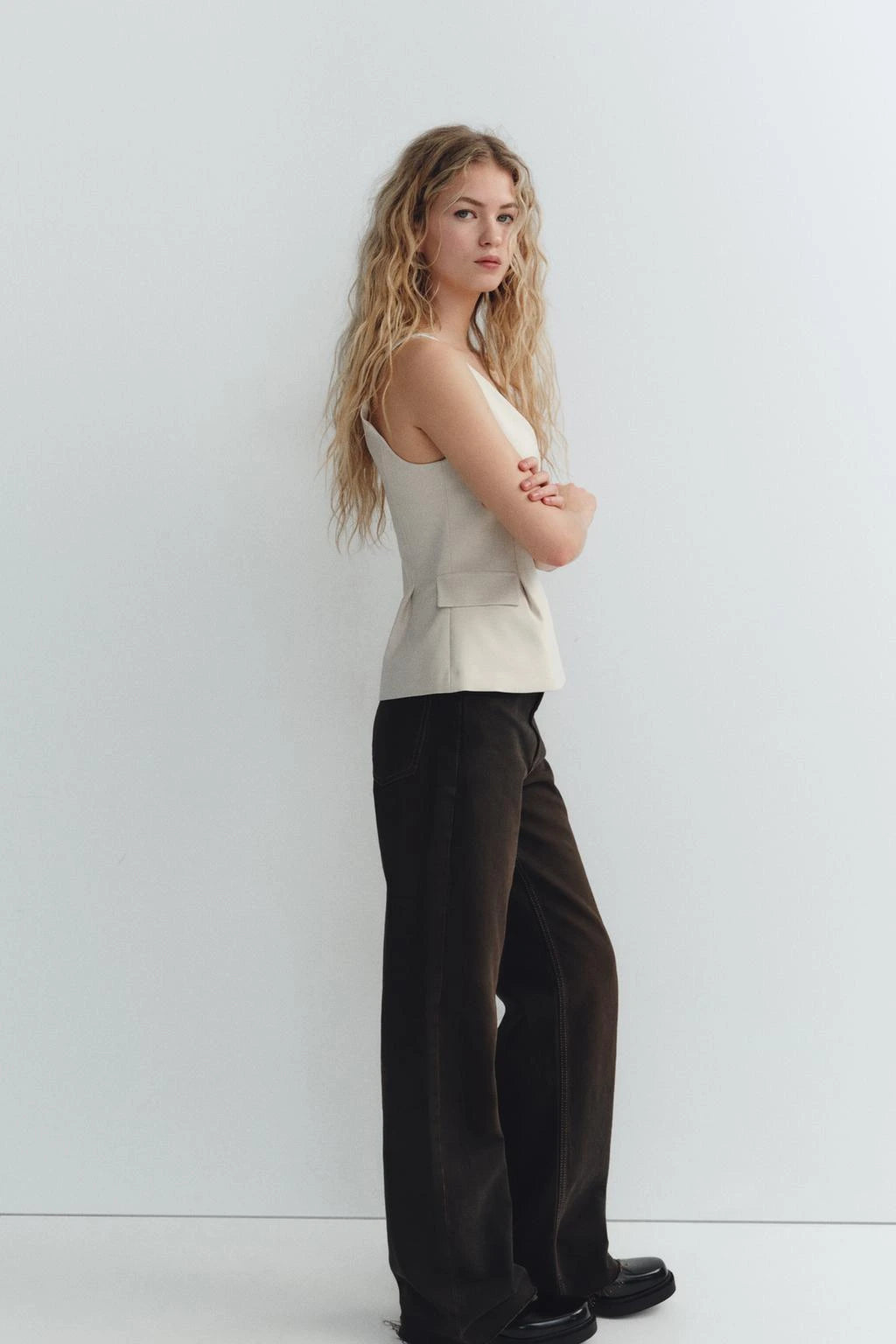 Woman wearing a beige sleeveless V-neck crop top with ruffled detail and zipper, styled casually in a café setting.