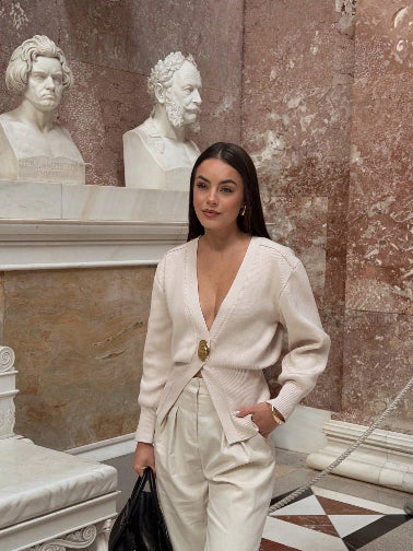 Model wearing a V-neck knit cardigan over a white tank top and trousers, enjoying a coffee outdoors in spring.