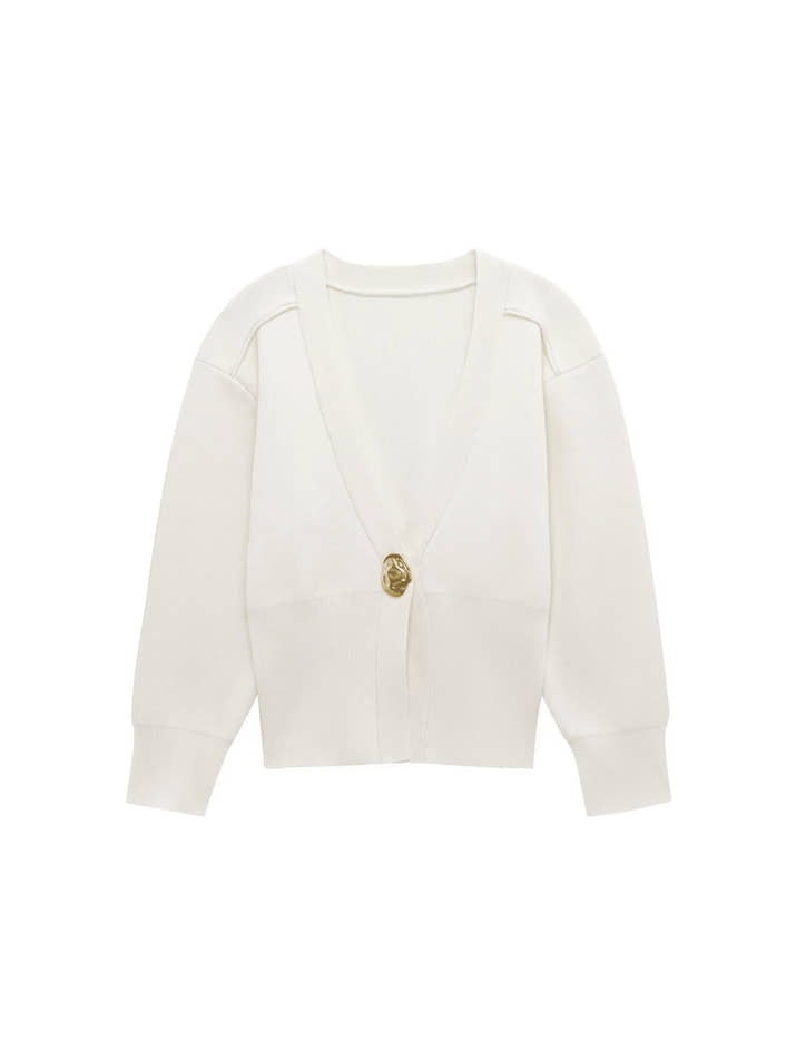 Model wearing a V-neck knit cardigan over a white tank top and trousers, enjoying a coffee outdoors in spring.