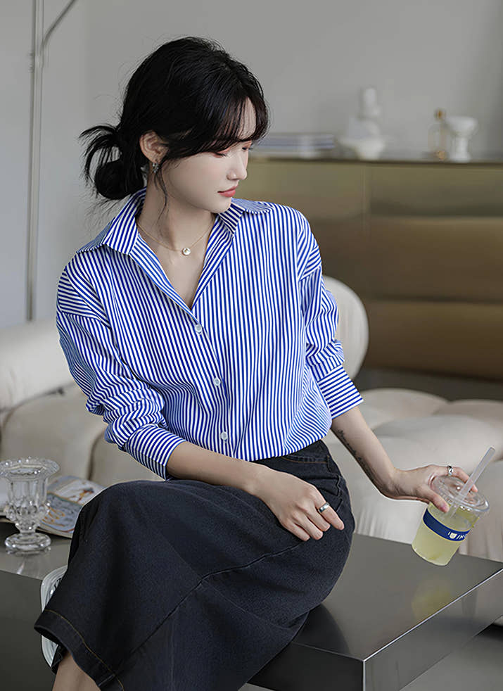 Woman wearing a blue-white striped loose office shirt with lantern sleeves in a minimalist urban setting.
