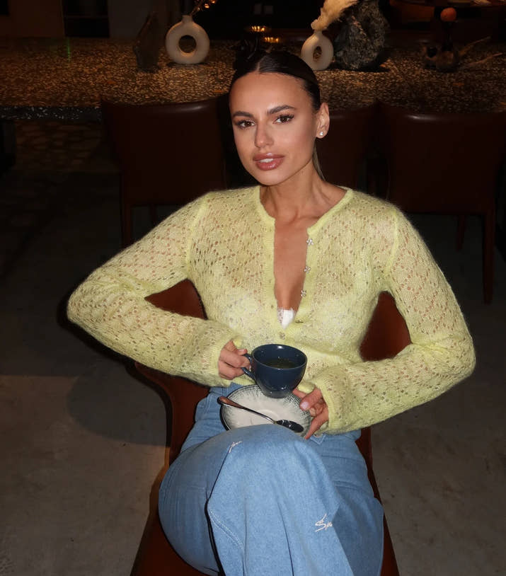 Woman in a yellow sweater holding a blue mug in a dimly lit room.