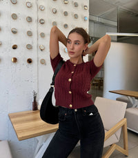 Woman posing in a modern indoor setting wearing a maroon knitted top and black jeans.