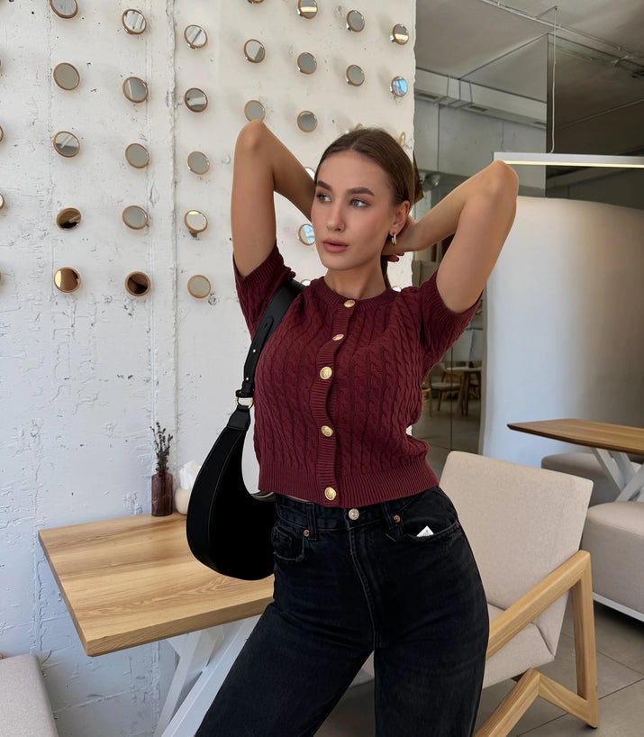 Woman posing in a modern indoor setting wearing a maroon knitted top and black jeans.