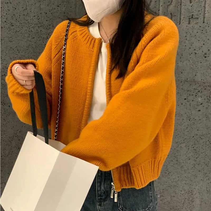 Person wearing a mustard yellow sweater holding a white bag against a gray wall.