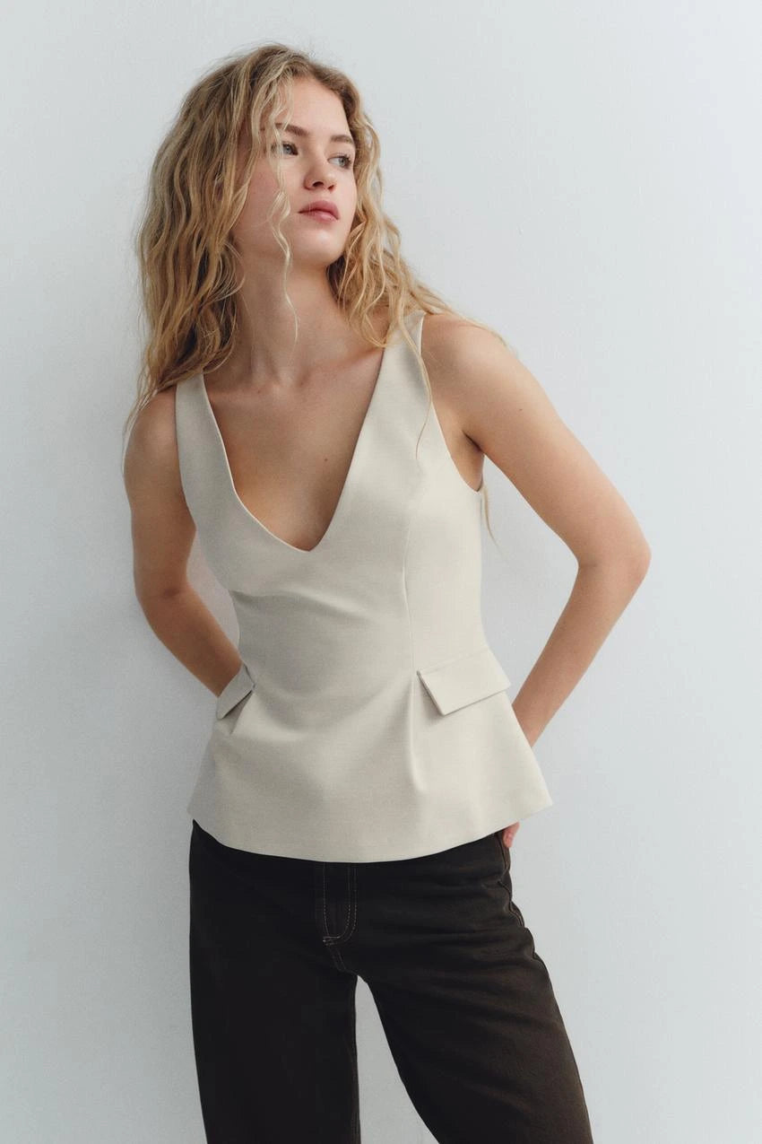 Woman wearing a beige sleeveless V-neck crop top with ruffled detail and zipper, styled casually in a café setting.