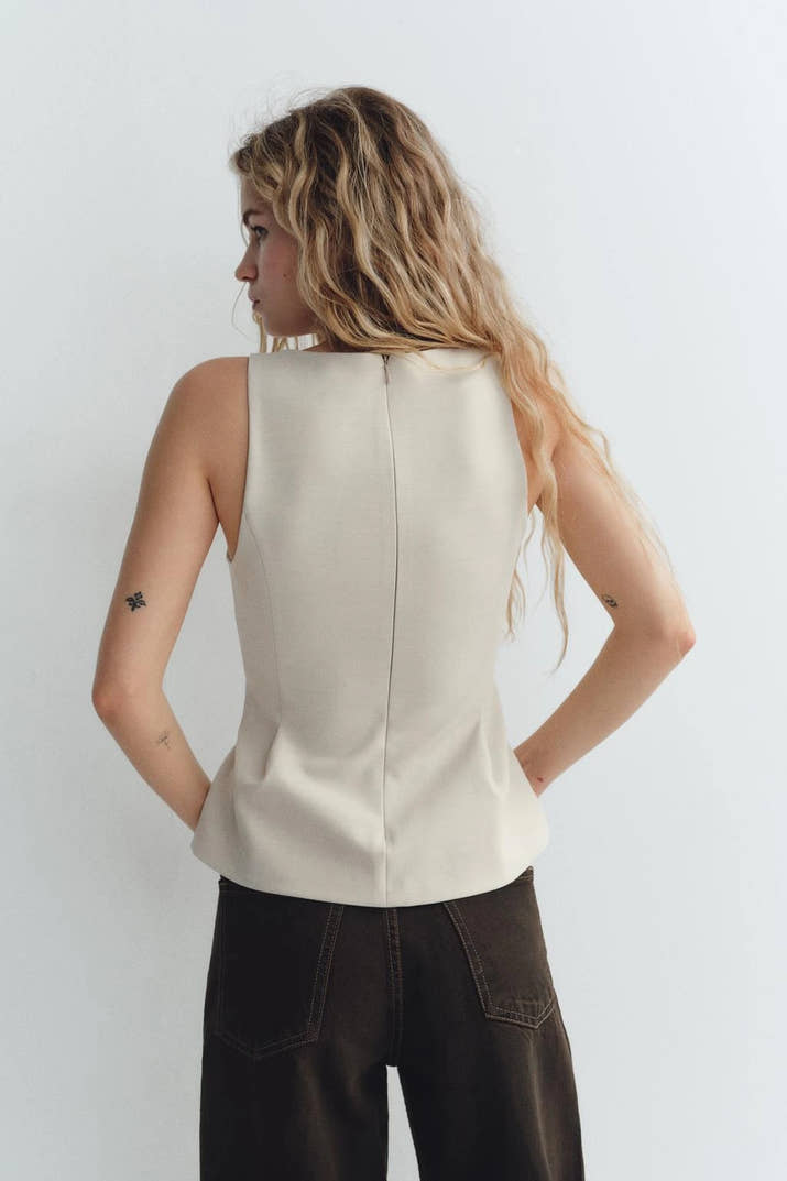 Woman wearing a beige sleeveless V-neck crop top with ruffled detail and zipper, styled casually in a café setting.