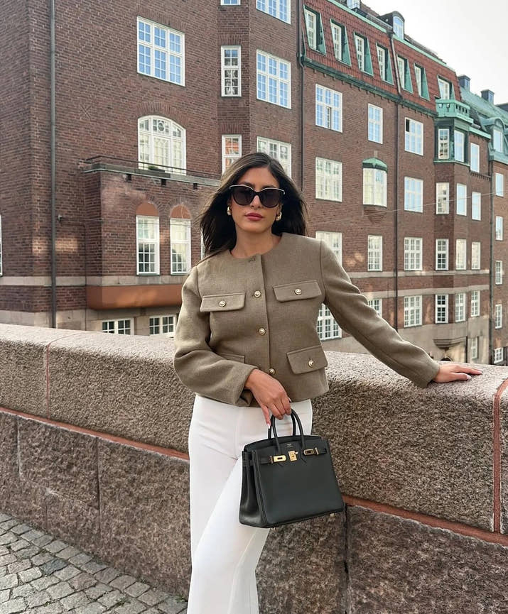 The woman looks casual and chic while walking around the city in a lightly cut khaki double-breasted tweed jacket with buttons.