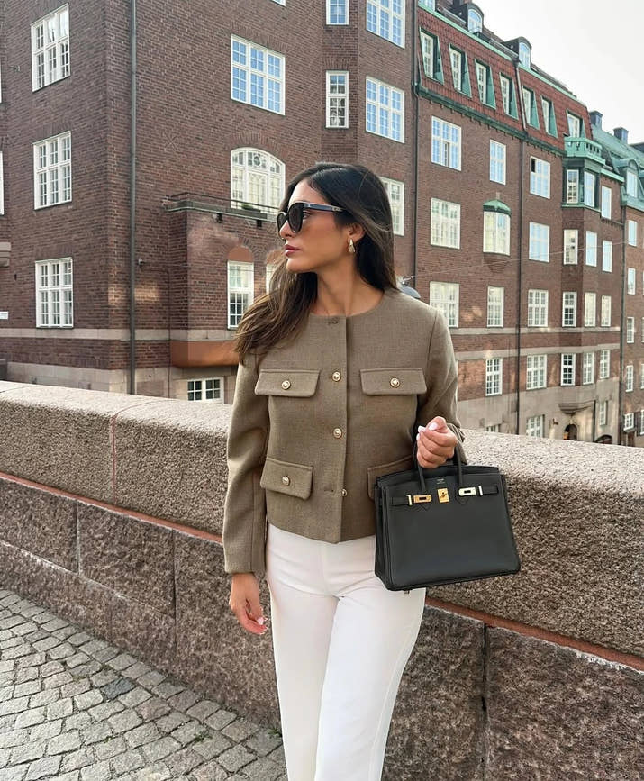 The woman looks casual and chic while walking around the city in a lightly cut khaki double-breasted tweed jacket with buttons.