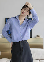 Woman wearing a blue-white striped loose office shirt with lantern sleeves in a minimalist urban setting.