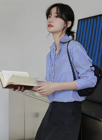 Woman wearing a blue-white striped loose office shirt with lantern sleeves in a minimalist urban setting.