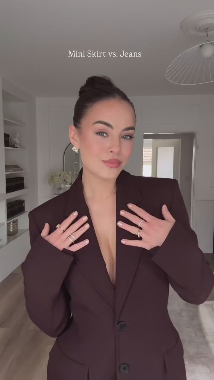 Woman wearing a slim-fit brown blazer jacket and A-line mini skirt in an office setting, confidently presenting in a modern workspace.